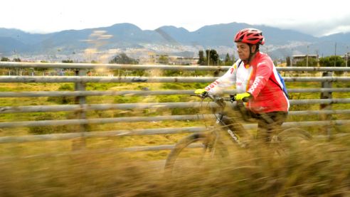 Mujer andando en bicicleta con casco e implementos protectores por un campo urbano.