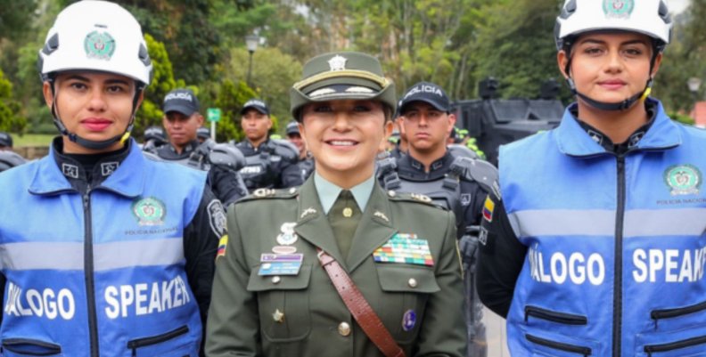 Mujer vestida de policía en el centro con dos mujeres con chalecos azules y cascos blancos a los lados