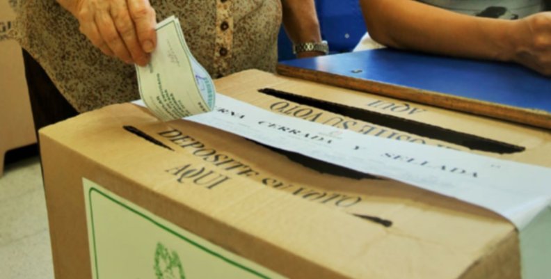 Urna de votación Una caja que funge como urna de votación con una ranura encima por la cual una mano va a introducir un papel de voto.