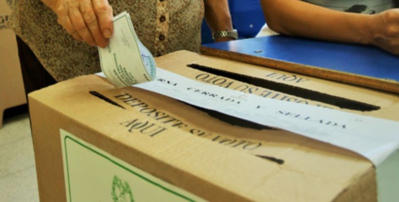 Una caja que funge como urna de votación con una ranura encima por la cual una mano va a introducir un papel de voto.