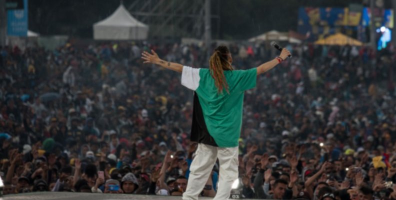 Hombre en el escenario con camisa verde y pantalón blanco abre los brazos frente al público