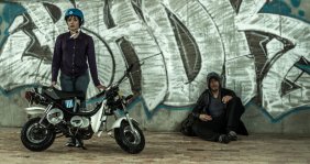 Dos personas, una mujer con casco y moto, un hombre sentado contra una pared.