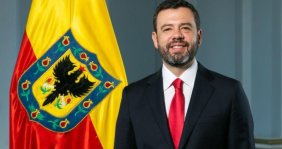 Hombre sonriente con traje negro, camisa blanca y corbata roja, delante de un fondo gris, y a su izquierda una bandera roja y amarilla
